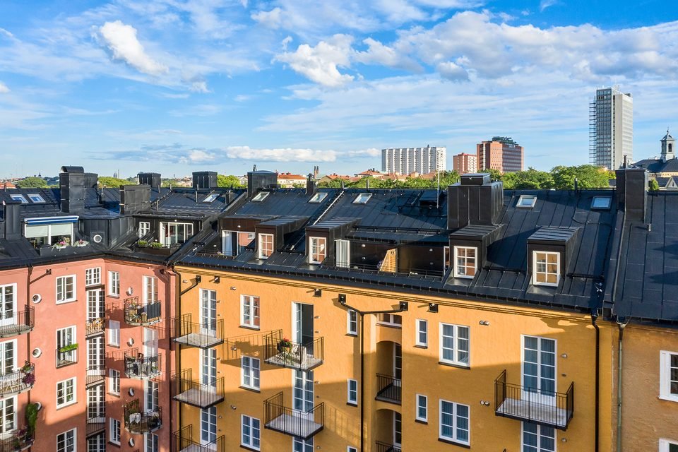 Husets plåttak med takkupor sett ovanifrån, med grönska och Stockholms stadssiluett bortom taknocken.