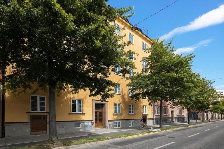 Husets gula fasad längs gatan, med en rad lövträd framför entrésidan.
